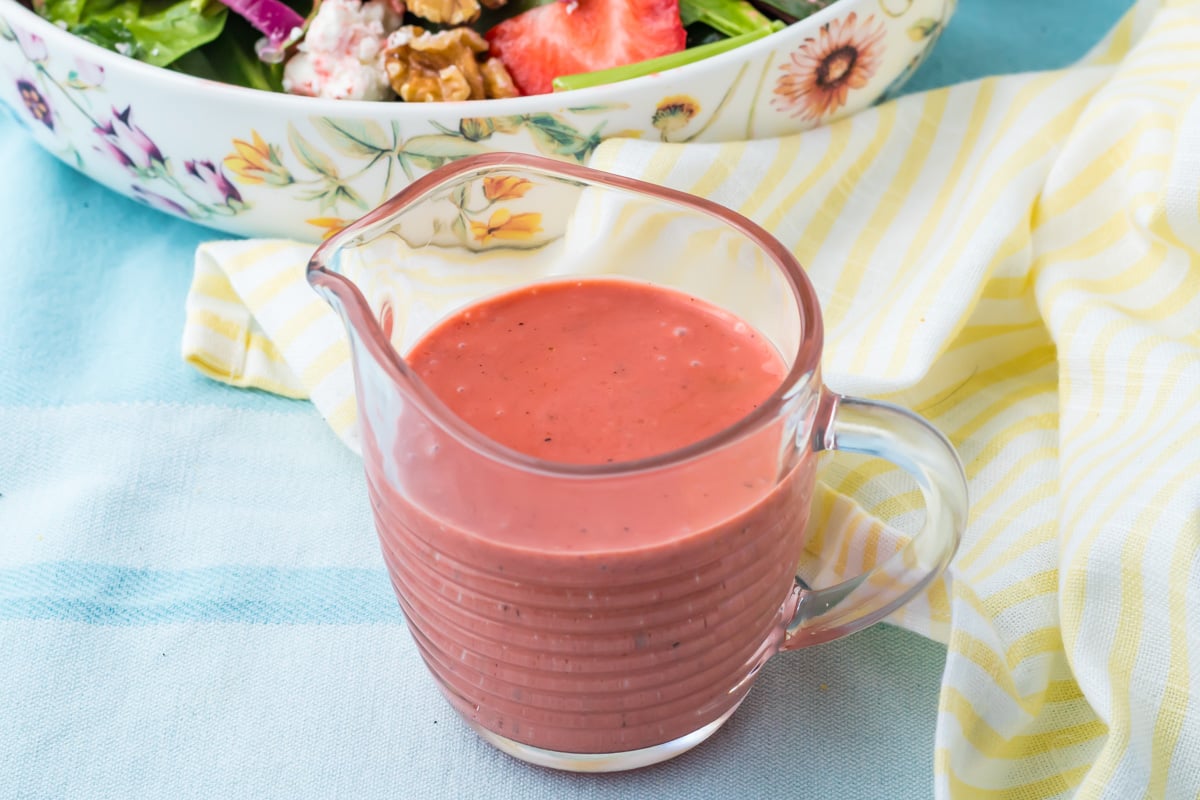 A glass pitcher filled with strawberry vinaigrette sits on a yellow-striped cloth, with part of a floral bowl of salad visible in the background.