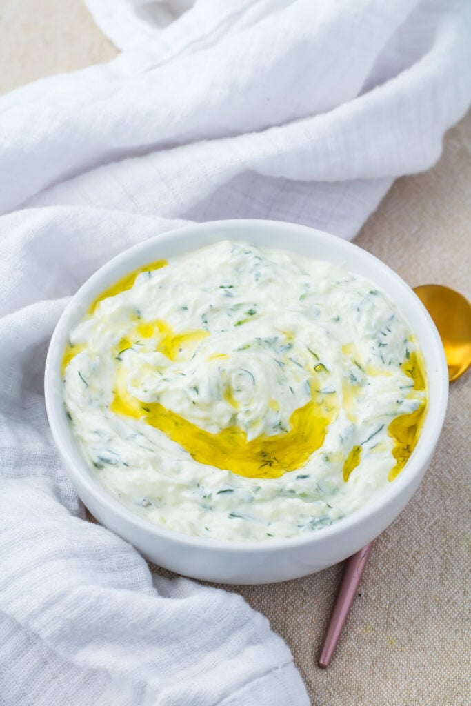A bowl of creamy tzatziki sauce drizzled with olive oil sits on a light cloth next to a gold spoon.