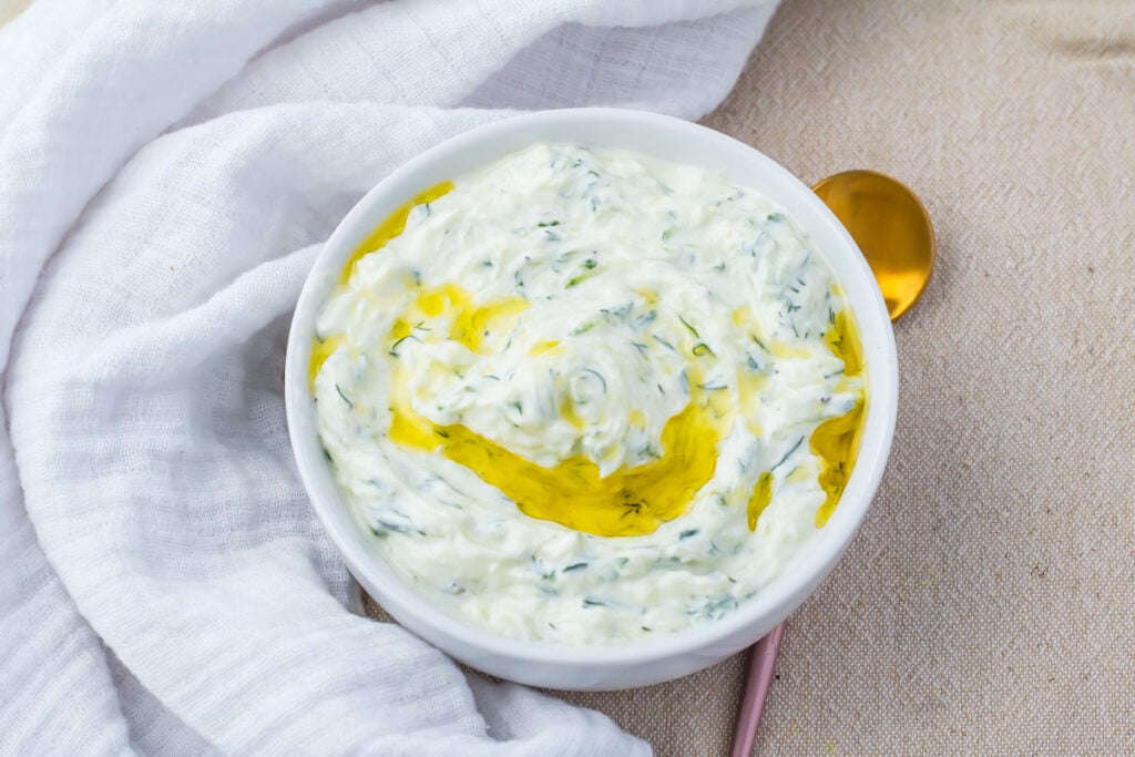 A white bowl of tzatziki dip with olive oil on top, placed on a beige surface next to a gold spoon and a white cloth.