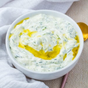 A bowl of creamy tzatziki sauce garnished with a swirl of olive oil, placed on a light-colored surface with a gold spoon and white cloth nearby.