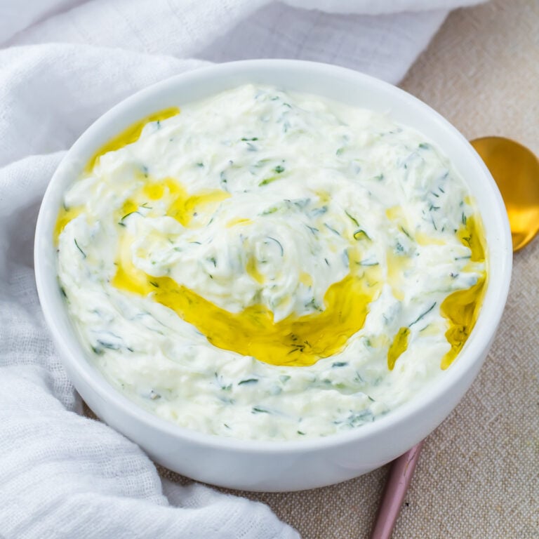 A bowl of creamy tzatziki sauce garnished with a swirl of olive oil, placed on a light-colored surface with a gold spoon and white cloth nearby.
