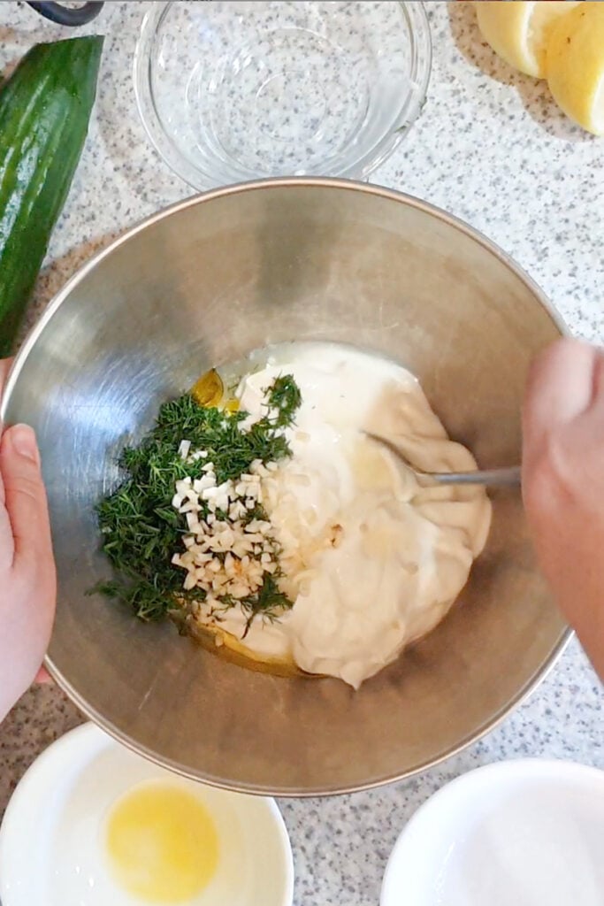 A person mixes yogurt, chopped garlic, dill, and olive oil in a metal bowl on a speckled countertop, with lemon, cucumber, and small bowls nearby.