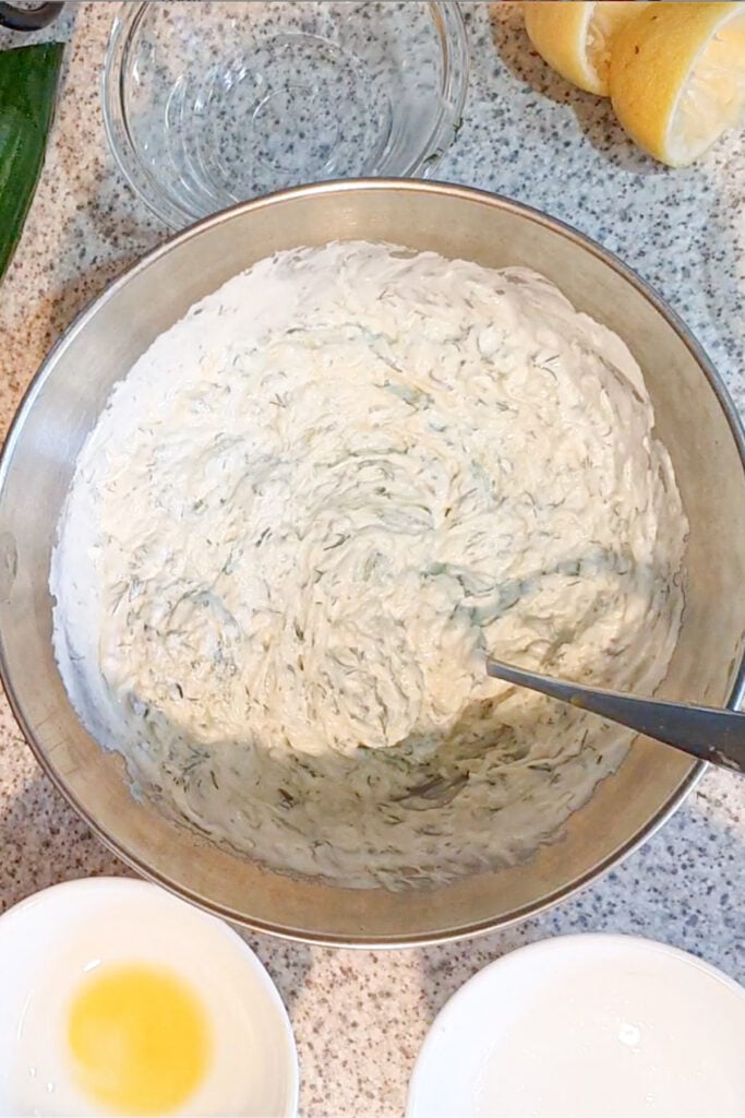 A metal bowl filled with a creamy, herbed mixture sits on a countertop next to a spoon, lemon halves, and small glass bowls containing oil and lemon juice.