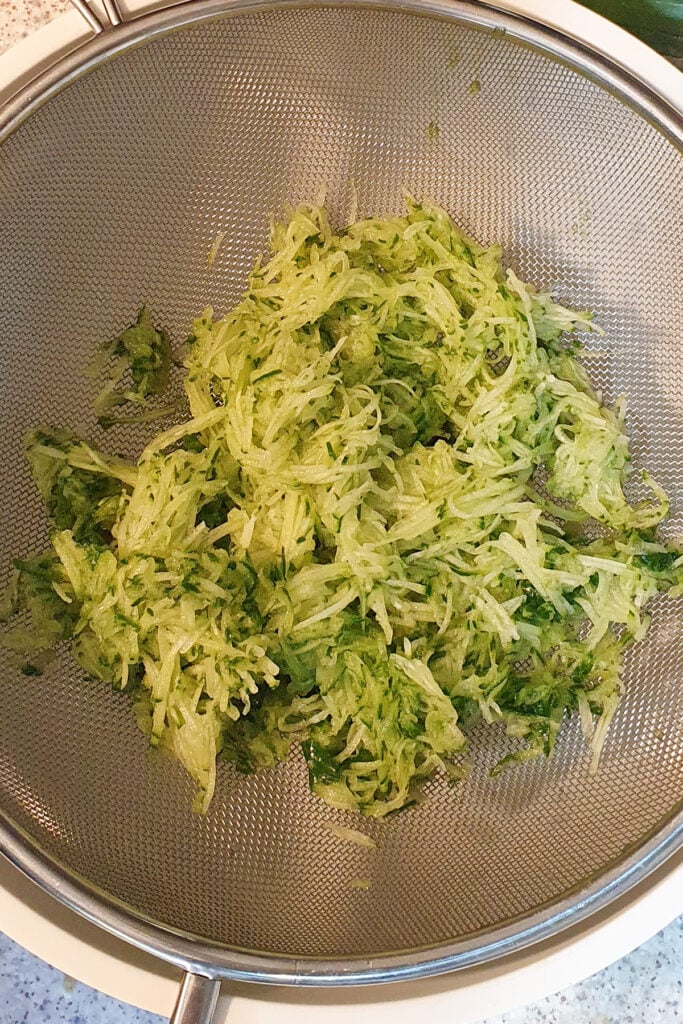 Shredded cucumber sits in a metal mesh strainer over a large bowl, with some liquid visible below.