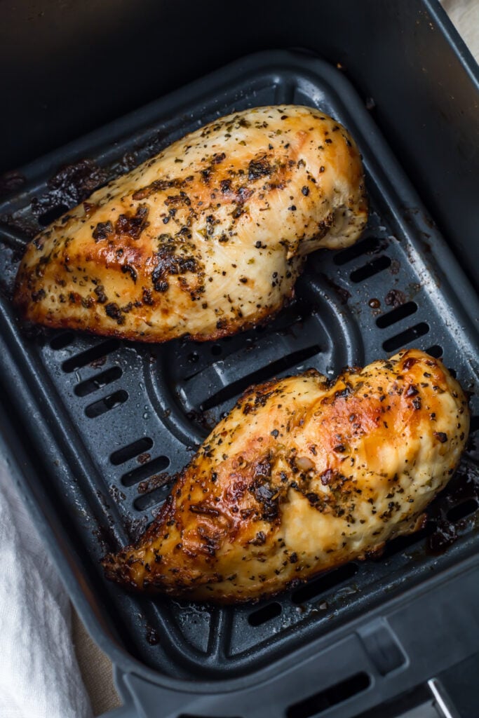 Two seasoned, cooked chicken breasts in a ninja air fryer basket, with browned tops and visible herbs on the surface.