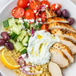 A Greek Chicken Grain Bowl with sliced grilled chicken, cherry tomatoes, cucumbers, kalamata olives, red onion, feta cheese, tzatziki, lemon wedge, and bulgur.