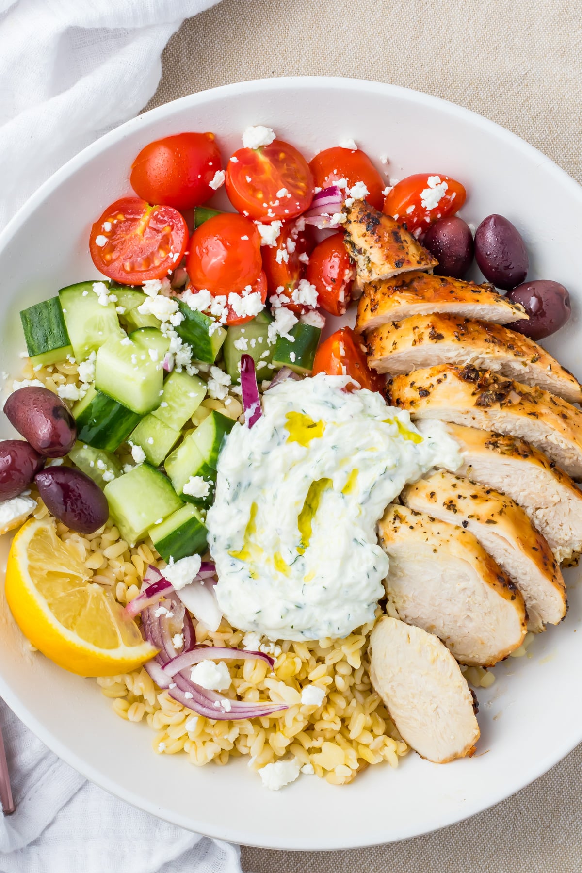 A Greek Chicken Grain Bowl with sliced grilled chicken, cherry tomatoes, cucumbers, kalamata olives, red onion, feta cheese, tzatziki, lemon wedge, and bulgur.