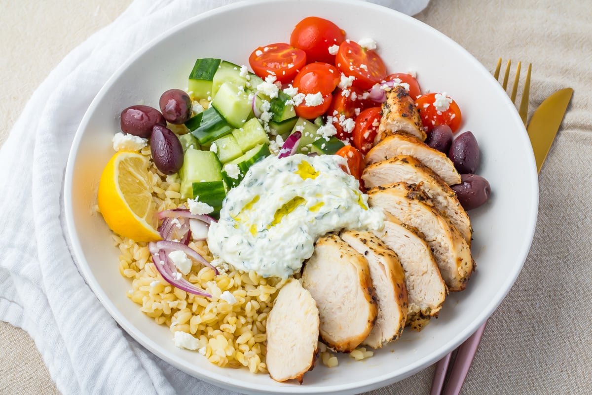 A Greek Chicken Grain Bowl with sliced grilled chicken, cherry tomatoes, cucumbers, kalamata olives, red onions, bulgur, tzatziki sauce, feta cheese, and a lemon wedge.
