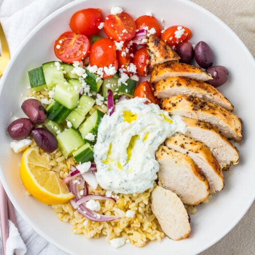 A Greek Chicken Grain Bowl with sliced grilled chicken, bulgur, cucumber, cherry tomatoes, Kalamata olives, red onion, feta cheese, tzatziki, bulgur wheat, and a lemon wedge.