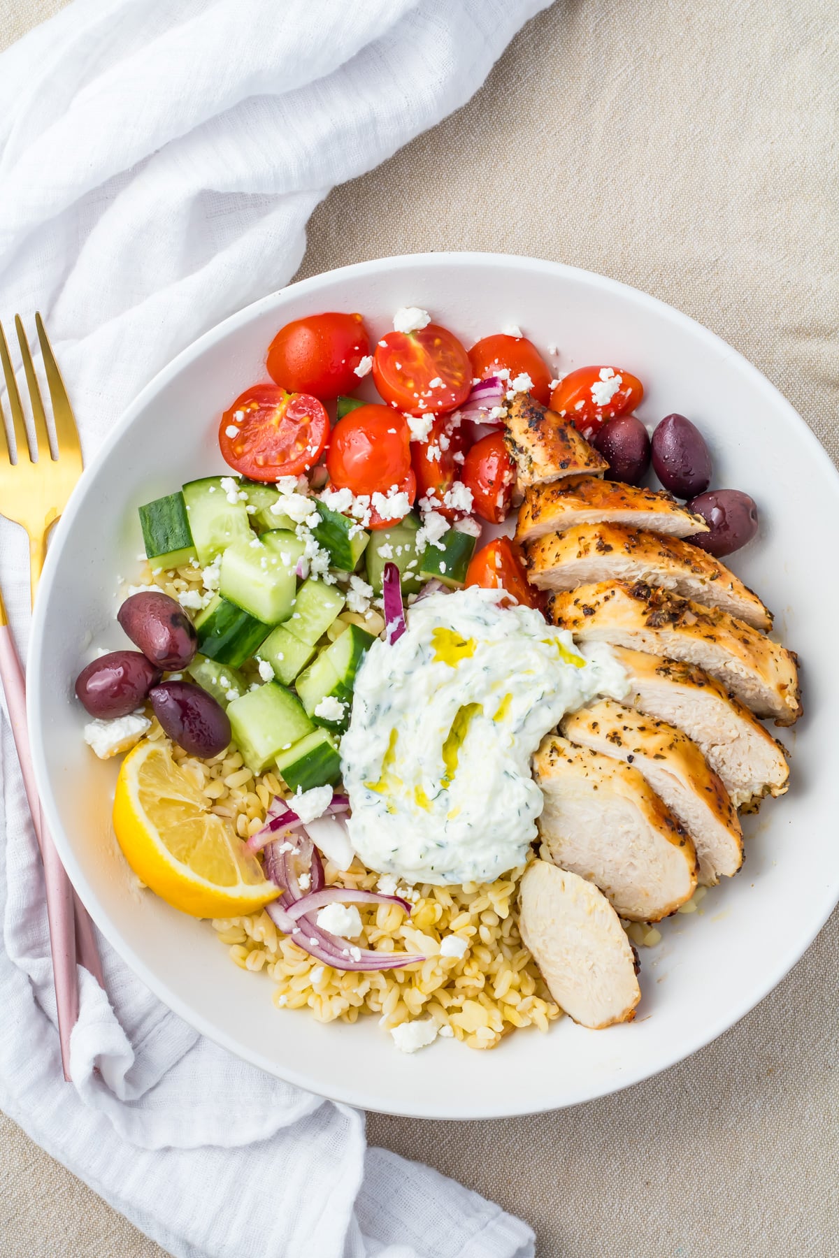 A Greek Chicken Grain Bowl with sliced grilled chicken, cherry tomatoes, cucumbers, Kalamata olives, red onions, feta cheese, tzatziki, bulgur, and a lemon wedge.