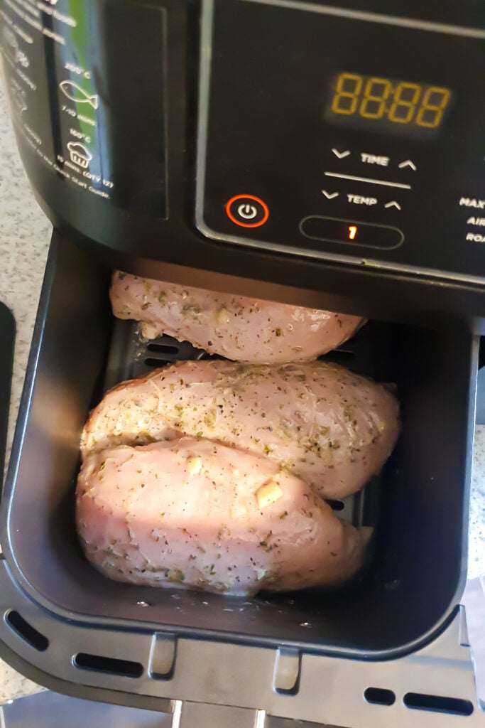 Three seasoned raw chicken breasts are placed inside an open Ninja air fryer basket, ready to be cooked. The air fryer control panel is visible above the basket.