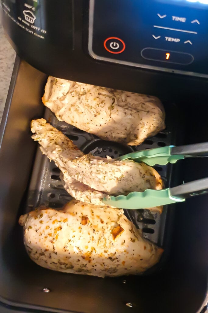 Three seasoned chicken breasts are being cooked in a Ninja air fryer; a pair of tongs is lifting one piece.