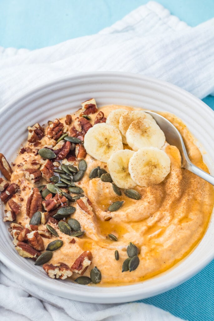 A bowl of creamy orange smoothie topped with banana slices, pecans, pumpkin seeds, and a sprinkle of cinnamon, with a spoon on the side.
