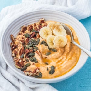 A pumpkin yogurt bowl topped with banana slices, pecans, pumpkin seeds, and cinnamon, on a white cloth with a spoon.