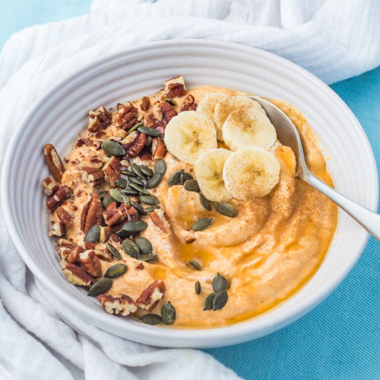 A pumpkin yogurt bowl topped with banana slices, pecans, pumpkin seeds, and cinnamon, on a white cloth with a spoon.