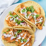 Three shredded slow cooker chicken tacos topped with chopped red onion, cilantro, and a drizzle of white sauce on a white plate with a blue tablecloth and white napkin in the background.