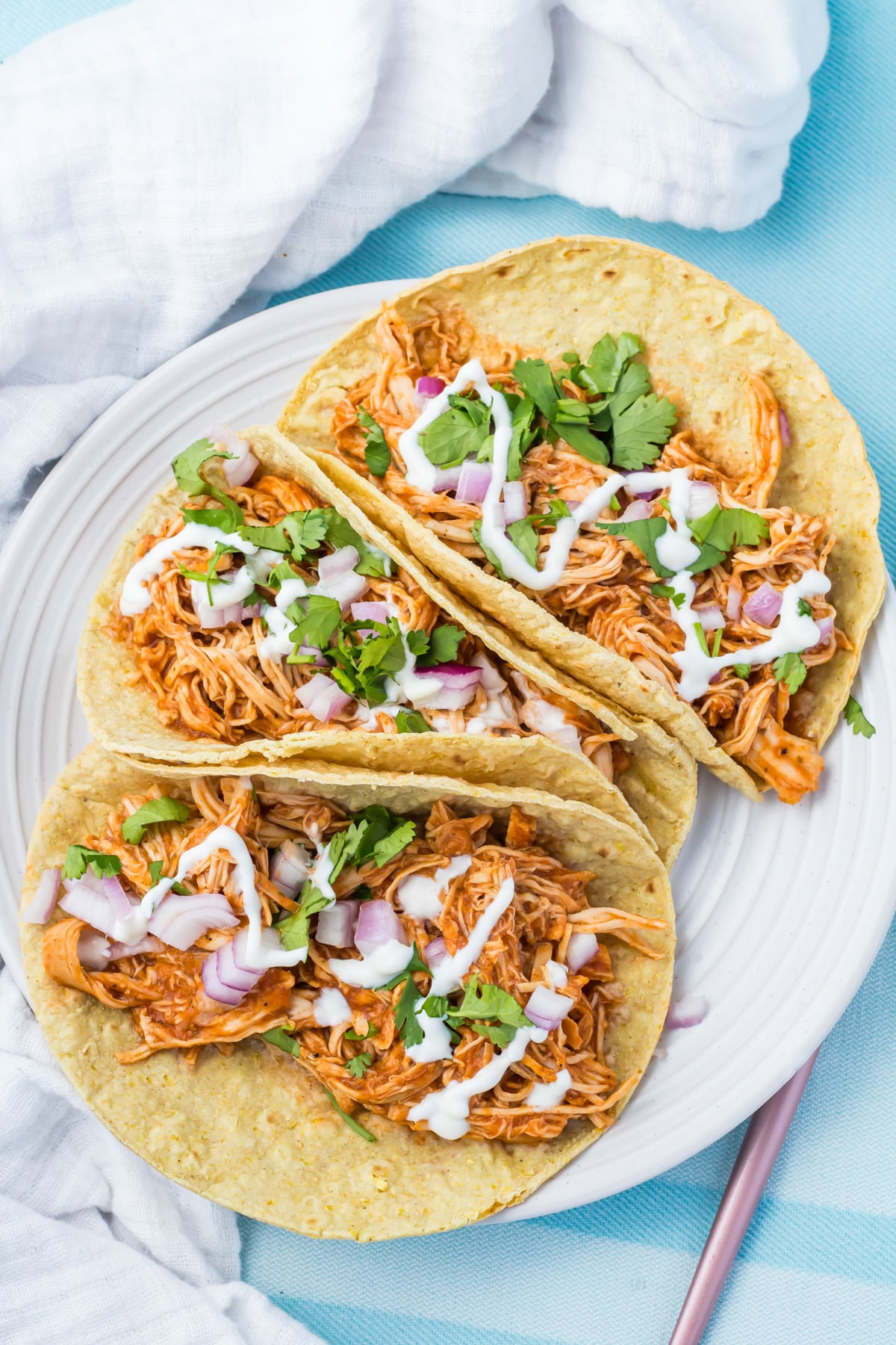Three shredded slow cooker chicken tacos topped with chopped red onion, cilantro, and a drizzle of white sauce on a white plate with a blue tablecloth and white napkin in the background.