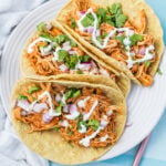 Three shredded slow cooker chicken tacos topped with chopped red onion, cilantro, and a drizzle of white sauce on a white plate.