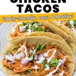 Three shredded chicken tacos topped with chopped red onion, cilantro, and a drizzle of white sauce on corn tortillas, served on a plate. Text overlay describes slow cooker chicken tacos.