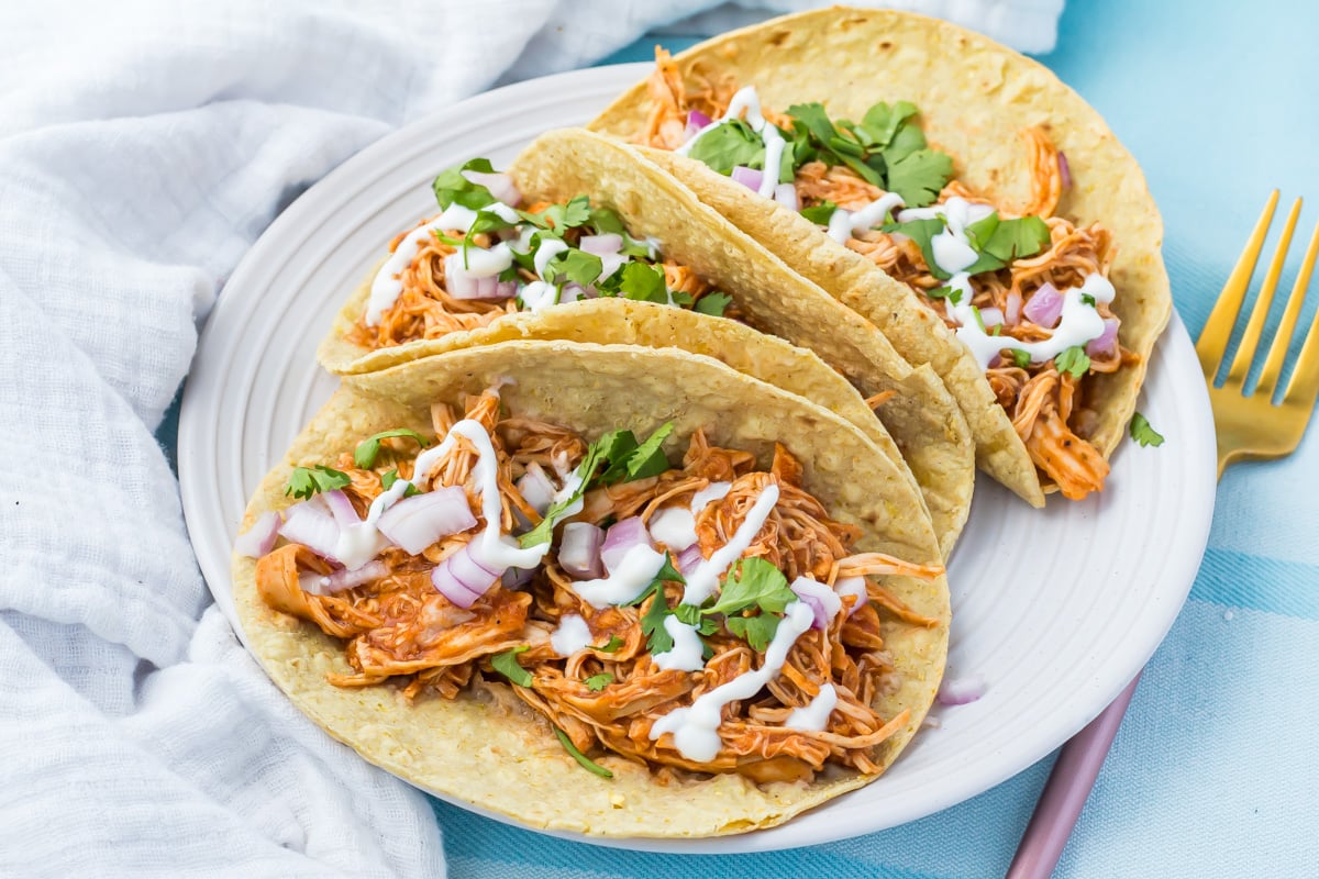 Three shredded slow cooker chicken tacos on corn tortillas topped with chopped red onions, cilantro, and a drizzle of white sauce, served on a white plate with gold utensils.