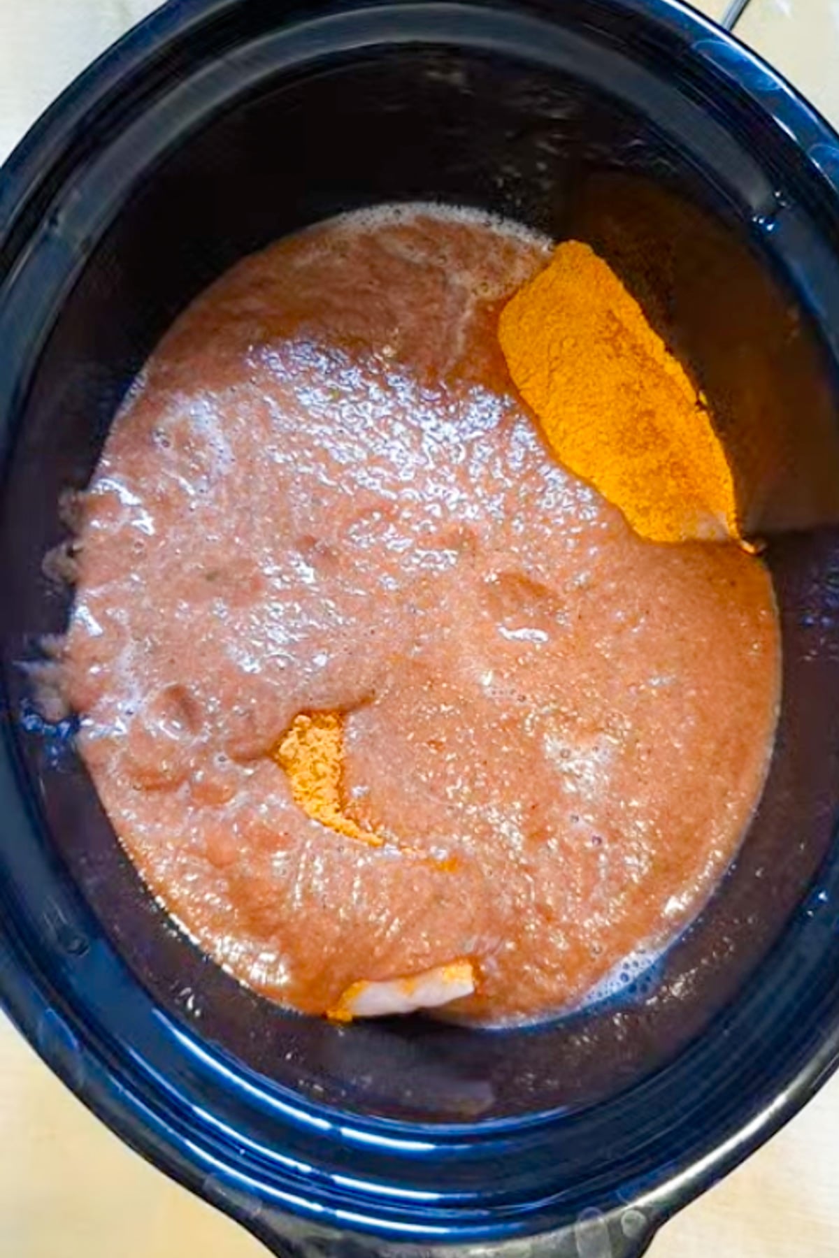 A slow cooker filled with a pinkish liquid mixture and visible clumps of yellow-orange powder on the surface.