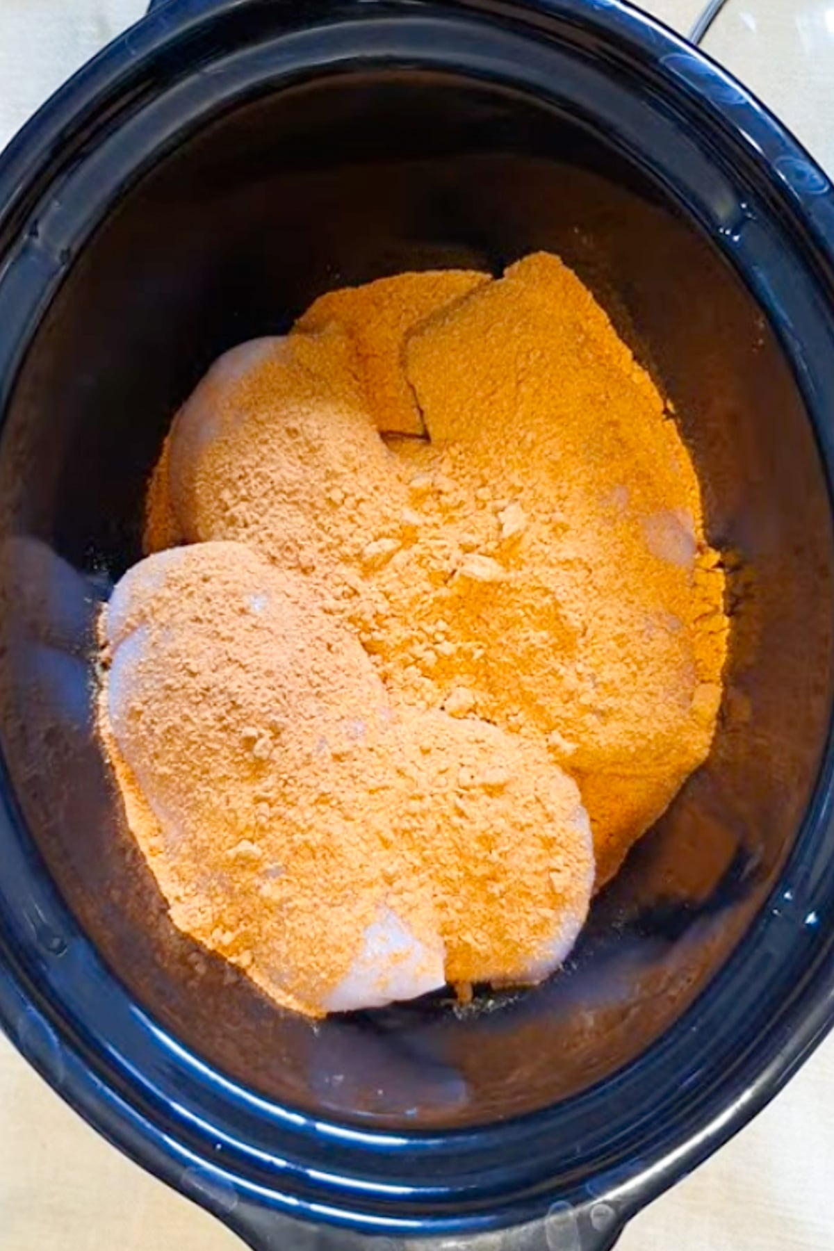 Raw chicken breasts in a slow cooker are covered with a layer of orange seasoning powder.