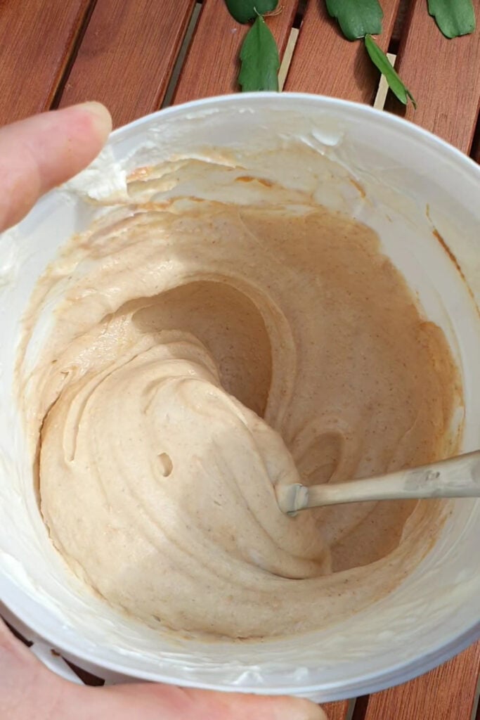 A hand holds a container of creamy beige mixture with a spoon inside, resting on a wooden table.