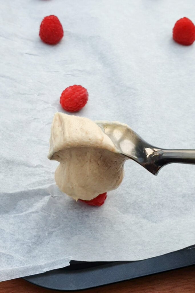 A spoon holds a dollop of cream above parchment paper scattered with raspberries.