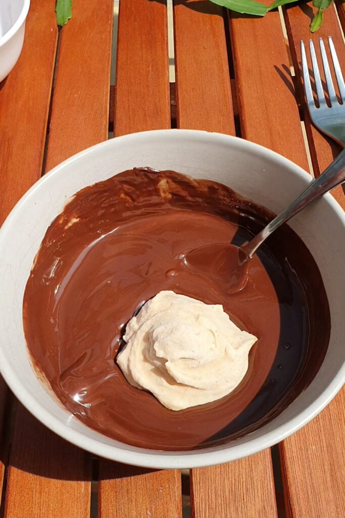 A bowl of melted chocolate with a dollop of whipped cream and a spoon, placed on a wooden table.