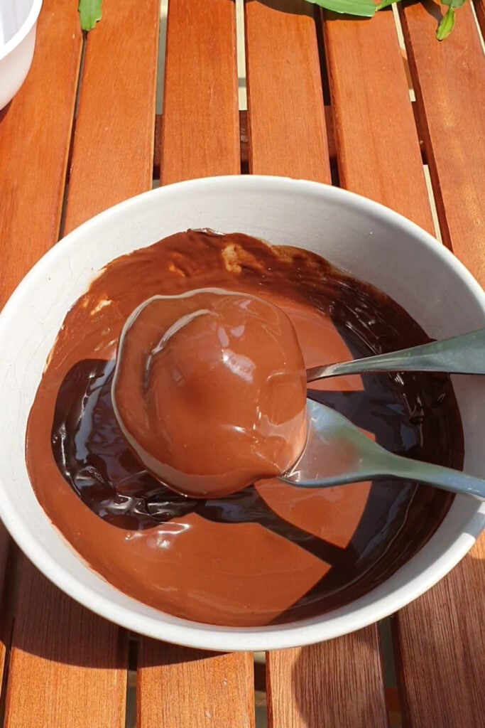 A white bowl with melted chocolate and two spoons holds a round dessert, on a wooden table.