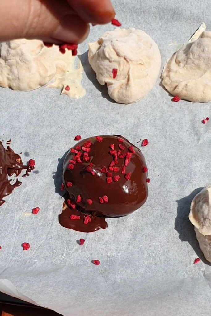 A chocolate-covered treat is sprinkled with small red pieces on parchment paper, surrounded by uncoated meringue cookies.