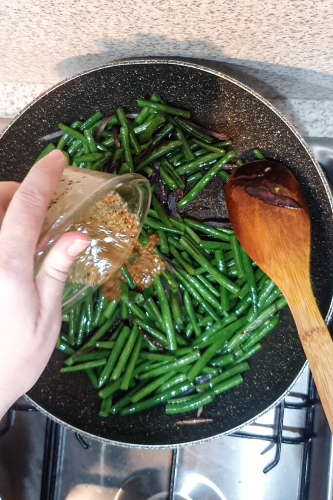 A hand pours a honey mustard seasoning mixture from a small glass bowl onto green beans cooking in a frying pan, next to a wooden spoon on a stovetop—perfect for delicious Honey Mustard Green Beans.