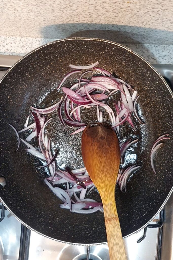 Sliced red onions being sautéed in a black frying pan with a wooden spoon on a stovetop, ready to be added to flavorful Honey Mustard Green Beans.