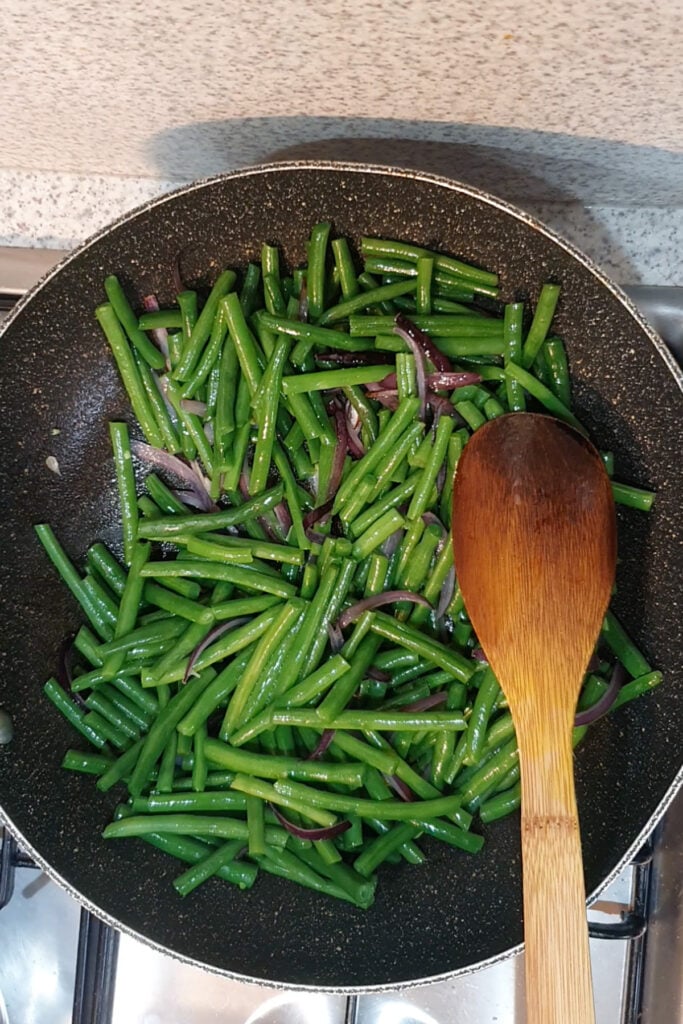Honey Mustard Green Beans and thinly sliced red onions are being sautéed in a nonstick pan with a wooden spoon on a stovetop.