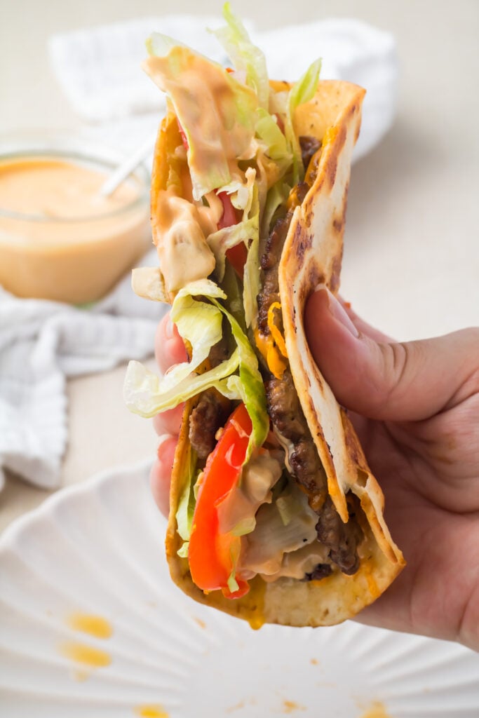 A hand holds a folded smash burger taco with seasoned beef, shredded lettuce, sliced tomatoes, cheese, and creamy sauce in a grilled tortilla.