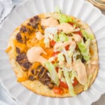 An open-faced smash burger taco on a white plate topped with ground beef, melted cheese, lettuce, tomato slices, and a creamy orange sauce.