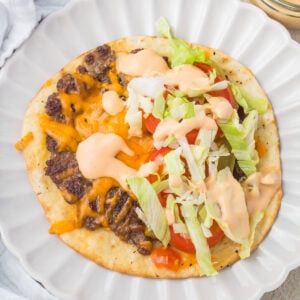An open-faced smash burger taco on a white plate topped with ground beef, melted cheese, lettuce, tomato slices, and a creamy orange sauce.