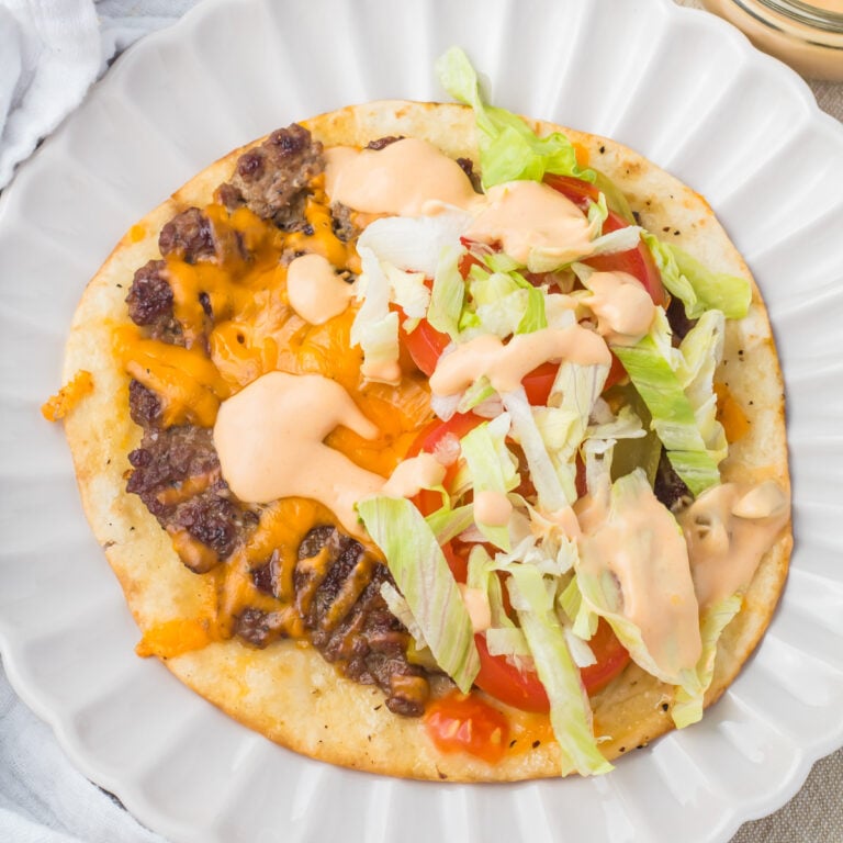 An open-faced smash burger taco on a white plate topped with ground beef, melted cheese, lettuce, tomato slices, and a creamy orange sauce.