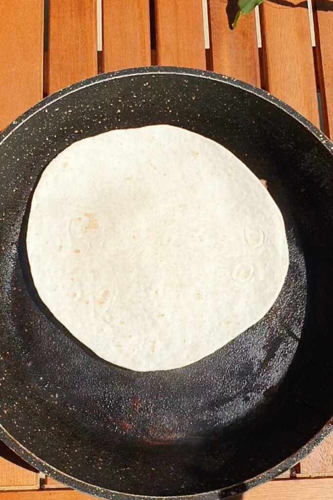 A plain flour tortilla is heating in a black frying pan placed on a wooden surface.
