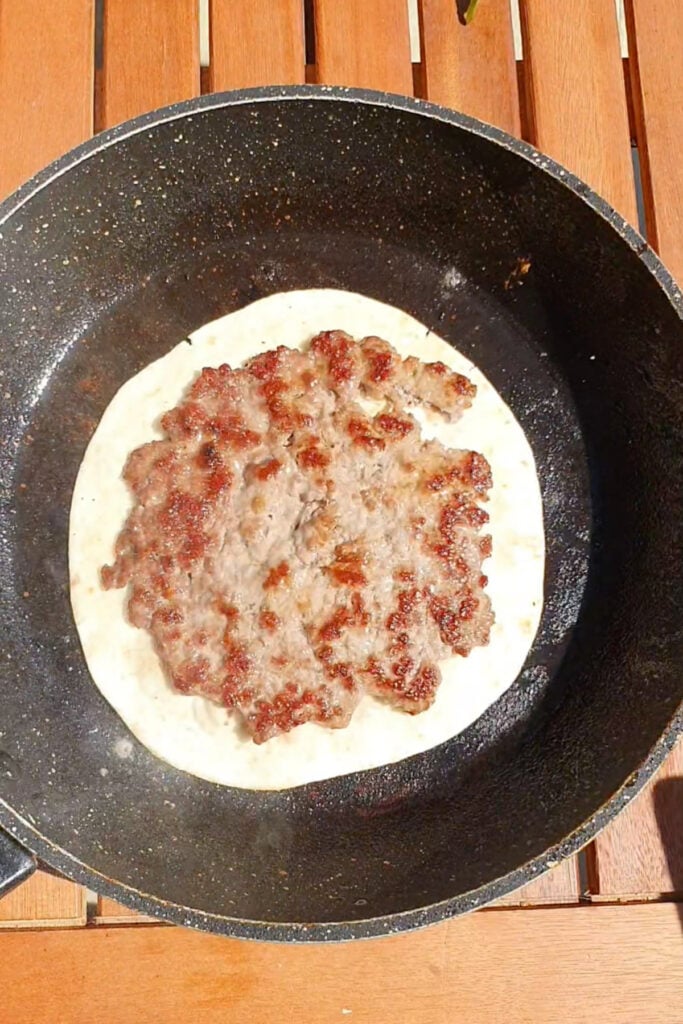 A cooked ground meat patty sits on a tortilla inside a nonstick frying pan on a wooden surface.