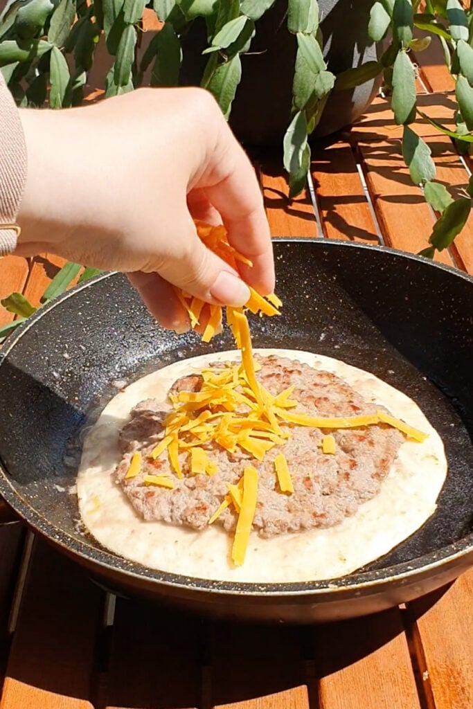 A hand sprinkles shredded cheddar cheese onto a cooked beef patty placed on a tortilla in a frying pan, with a plant and wooden surface in the background.