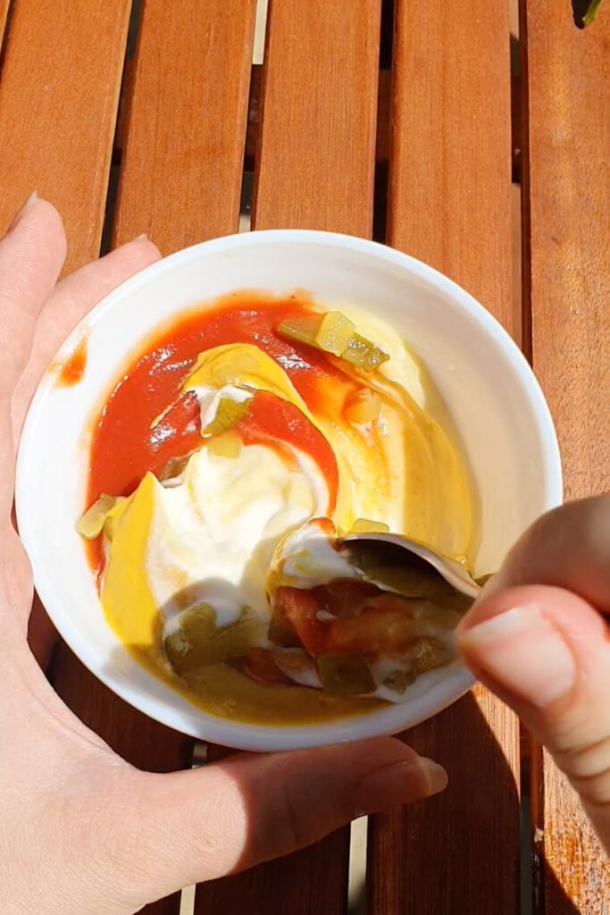 A hand holds a small white bowl with ketchup, mustard, mayonnaise, and chopped pickles being mixed with a spoon over a wooden table.