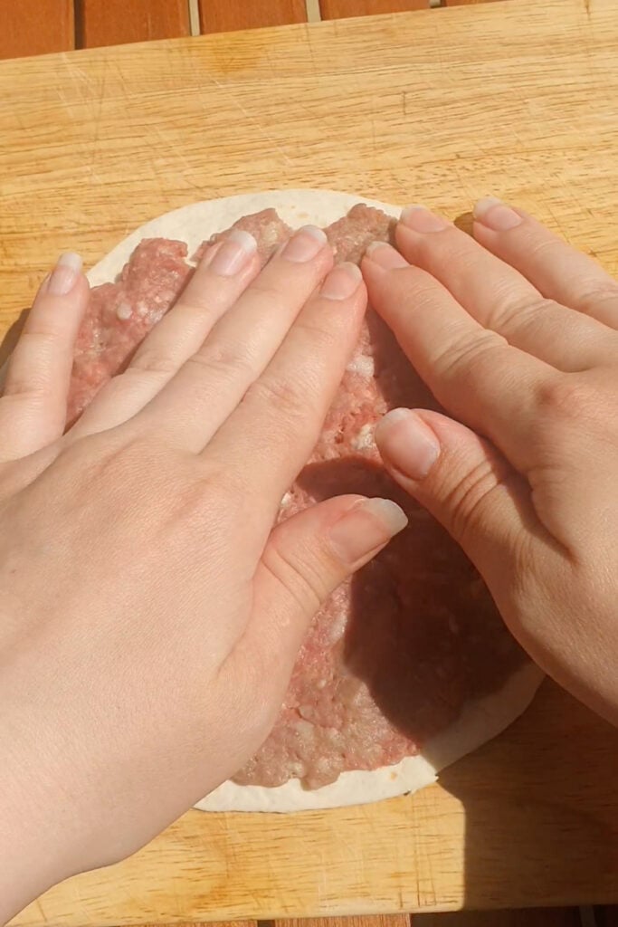Two hands pressing raw ground meat onto a round flatbread or dough on a wooden surface.