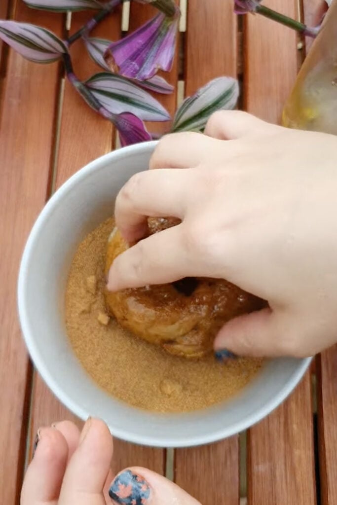A hand presses a donut into brown sugar in a white bowl on a wooden table, with some purple-striped leaves nearby.