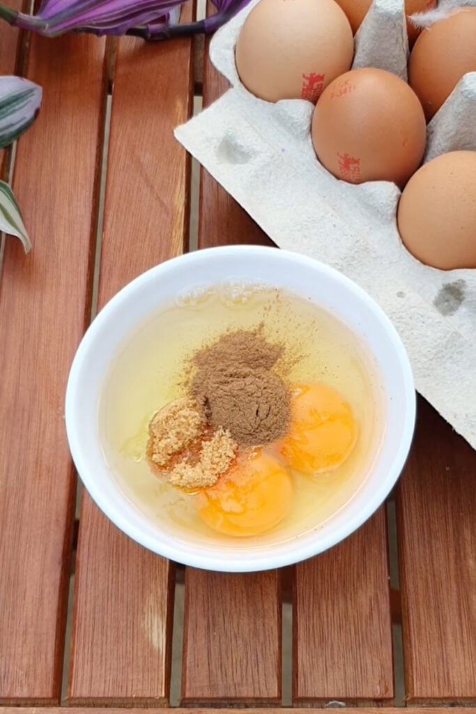 A white bowl with three raw eggs, brown sugar, and ground cinnamon next to a carton of eggs on a wooden table.