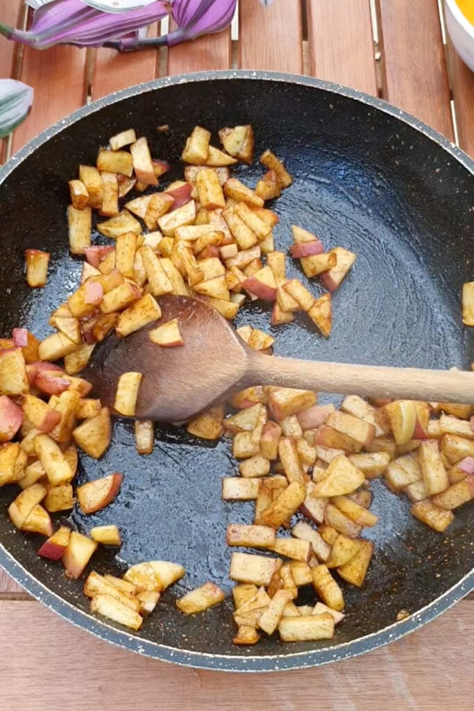 Diced apples with spices are being sautéed in a nonstick skillet with a wooden spoon on a wooden table.
