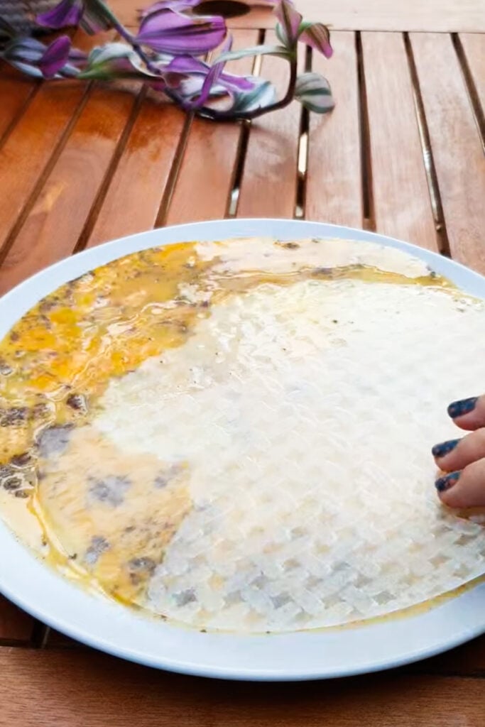 A hand presses a sheet of rice paper onto a plate covered with beaten eggs and seasoning on a wooden table.