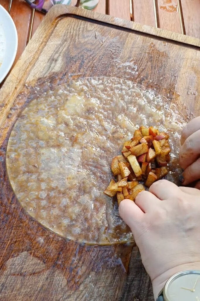 Hands wrapping a rice paper sheet around a filling of chopped, seasoned apples on a wooden cutting board.