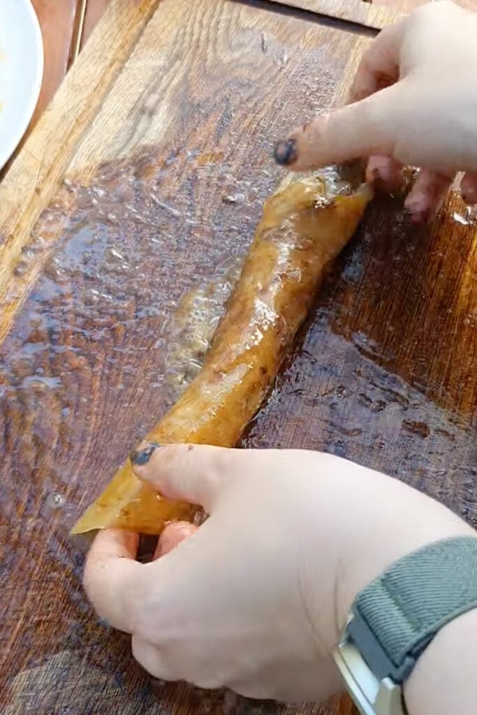 Hands rolling a translucent sheet of rice paper, on a wooden cutting board.