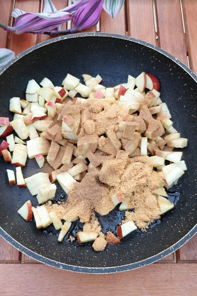 Diced apples with brown sugar and spices are cooking in a black frying pan on a wooden surface, with plant leaves visible in the background.